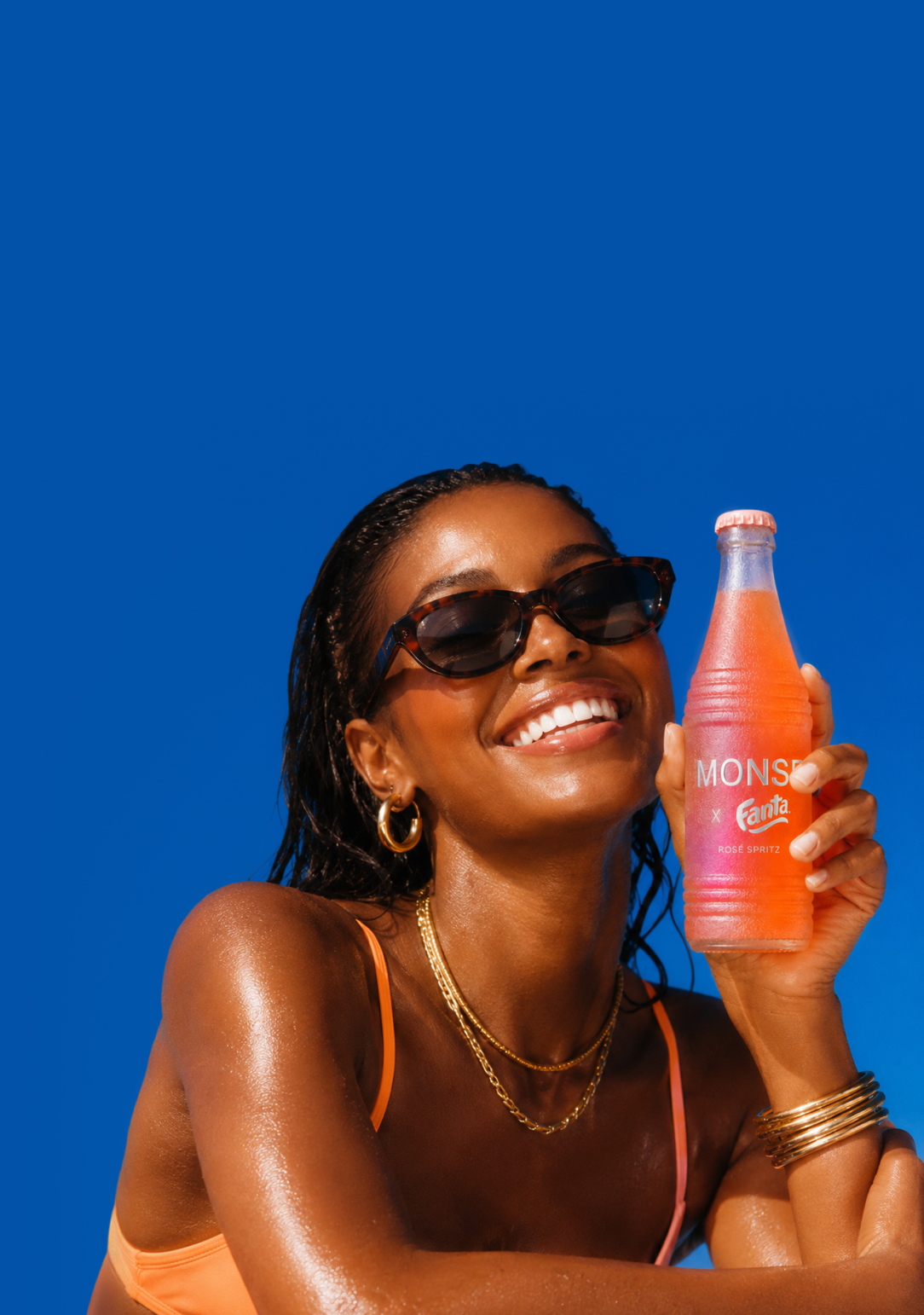A model laughing in the sun holding a MONSE x Fanta Rosé Spritz bottle against a cobalt blue sky